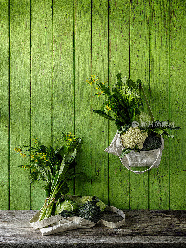 绿叶蔬菜挂在一个可重复使用的棉花袋从一个钉子，在一个旧的绿色木板墙的背景与其他蔬菜在一个可重复使用的袋子风化桌子下面。
