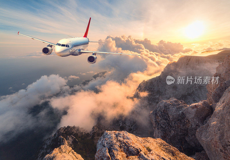 飞机在日落时飞过云层。白色的飞机，山脉，大海和橙色的天空和夏天的太阳。客机正在降落。商务旅行。商用飞机