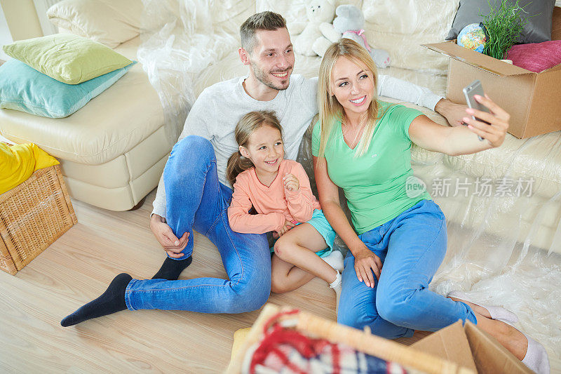 幸福美丽的年轻家庭在休闲服装坐在地板上，倚在聚乙烯沙发下，而摆姿势自拍在新公寓的智能手机