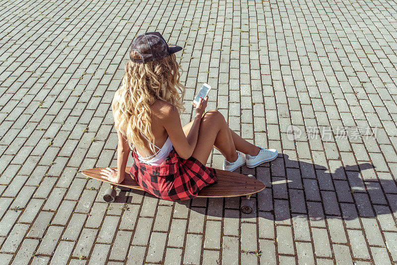 金发长头发的女孩，夏天的城市坐滑板，阅读信息应用在线，社交网络。时尚生活方式，现代理念概念潮流。晒黑的人物。背景铺地砖路。