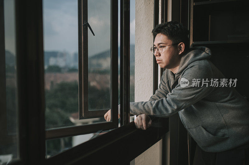 一个亚裔华人中年男子，穿着连帽衫，在日落时分从家里望向窗外，表情严肃