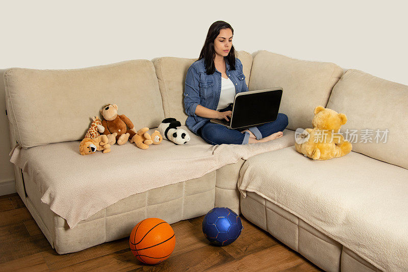 女人坐在沙发上，用笔记本电脑打字，周围有孩子们的玩具