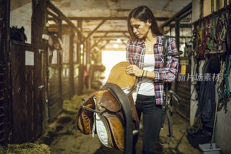 女人准备训练马，站在马厩里准备马鞍-一个年轻女人站在马厩里的马