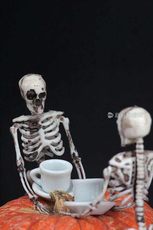 万圣节概念——两个骷髅在黑色背景下的南瓜桌上喝茶。垂直的图像