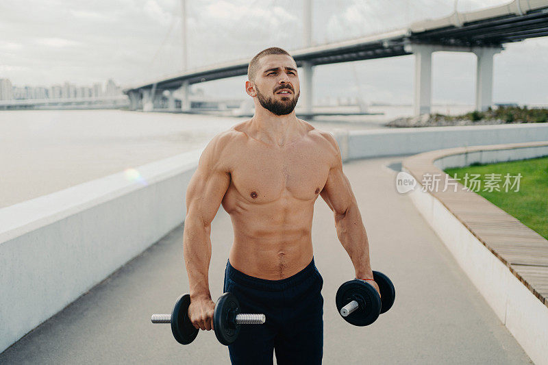 肌肉健美者集中到距离有强壮的身体强壮的肌肉拿杠铃和锻炼肱二头肌户外姿势附近的河与桥。健康的生活方式和运动理念