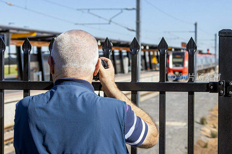 一名年长的男性火车观察员正在拍摄电动火车通过安全围栏的照片