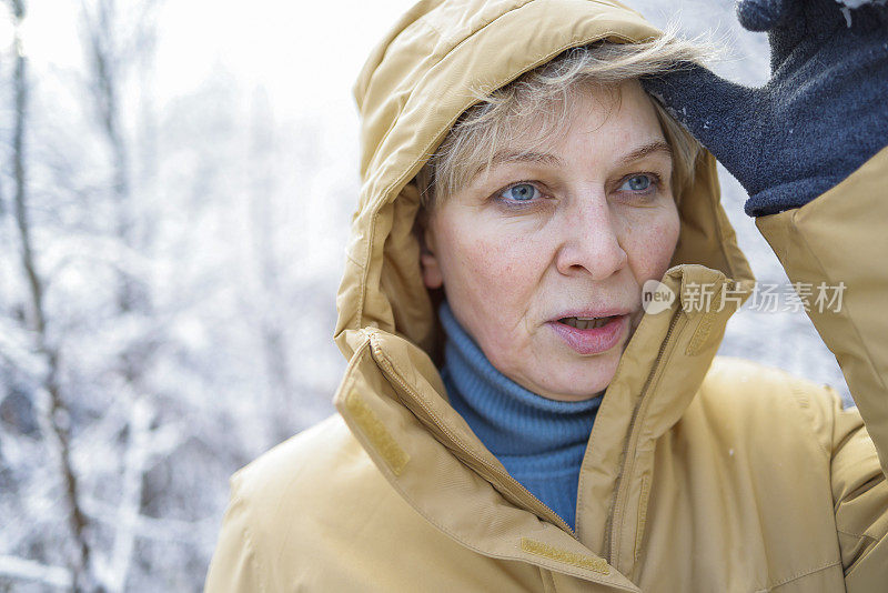 在下雪的公园里，一个成熟的女人穿着黄色的冬季夹克，头上戴着兜帽。