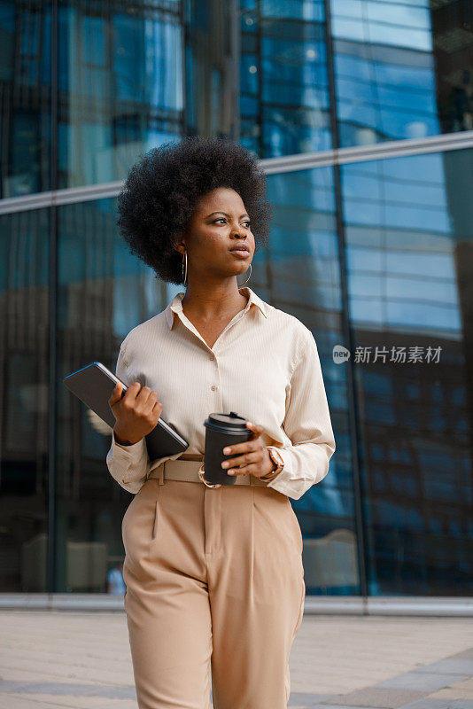 一位年轻漂亮的女商人，身着米色套装，端着一杯咖啡和一台数码平板电脑，走在商业区的街道上