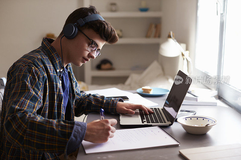 一个十几岁的男孩戴着耳机在卧室的书桌前工作