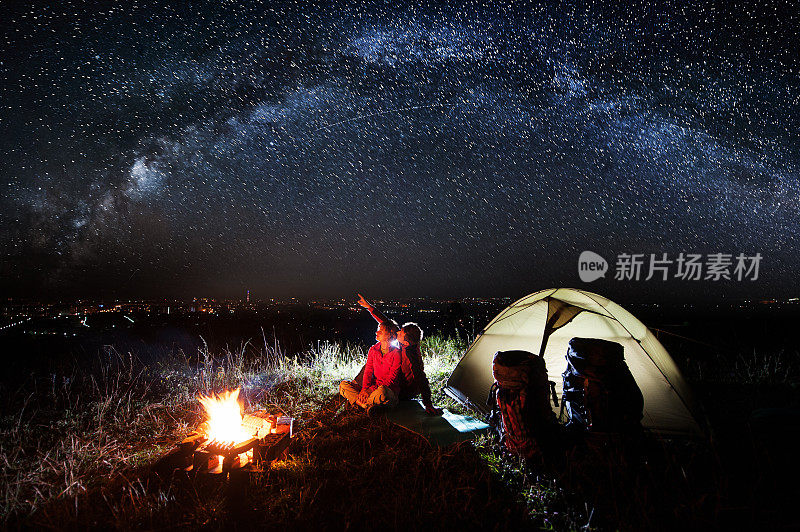 晚上在镇上附近露营。年轻的夫妇坐在篝火和帐篷附近，看着美丽的星空，欣赏夜景。盖伊指着天空。天体摄影术