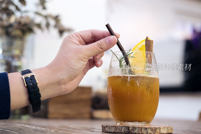 在咖啡馆的木桌上，一个女人的手拿着一根吸管在一杯橙冷煮咖啡