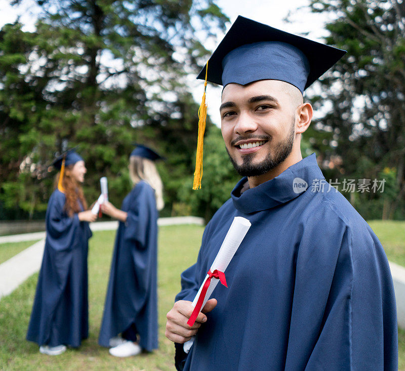 英俊的大学毕业生拿着他的文凭看着镜头微笑，而其他毕业生站在后面