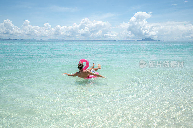 一个年轻人在田园般的海滩上放松，充气火烈鸟在清澈的海水中玩耍在泰国的岛屿。人们旅行奢侈，乐趣和酷的态度概念