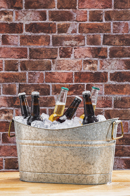 靠砖墙的木桌上放着一个镀锌的桶，上面放着各种各样的冰上啤酒瓶