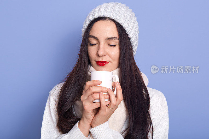 照片中，美丽的女孩端着一杯茶或咖啡，穿着暖和的毛衣和帽子，在蓝色的背景下摆出孤立的姿势，闭着眼睛站着，用一个盛着热饮的杯子晒手。