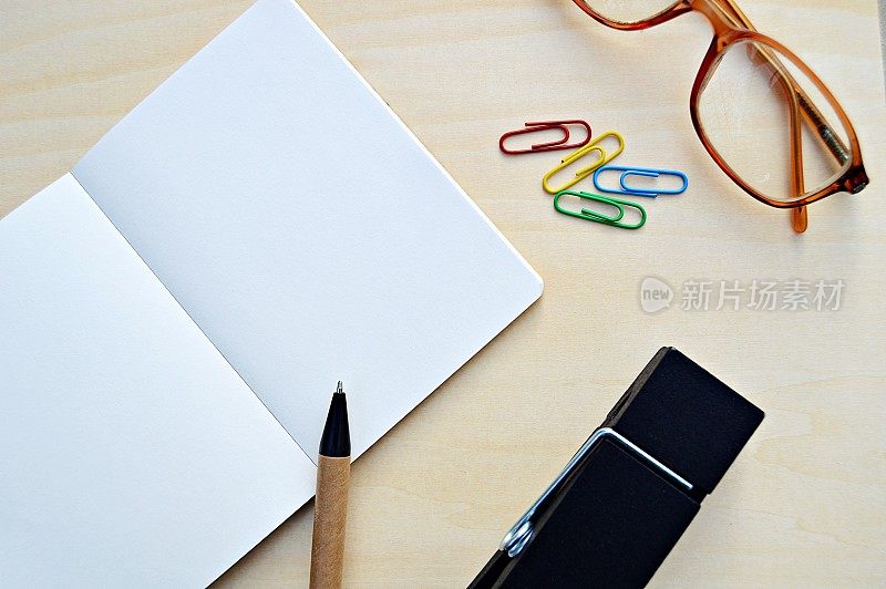 一张水平照片，一张空白笔记本的白色素色页，旁边是一支棕色和黑色的钢笔和彩色回形针，眼镜放在一个木制的米色水平背景上，很有美感。