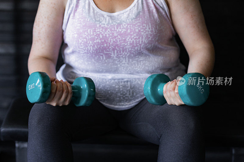 为减肥而进行的力量训练:一个匿名超重女性在健身房用哑铃锻炼的特写