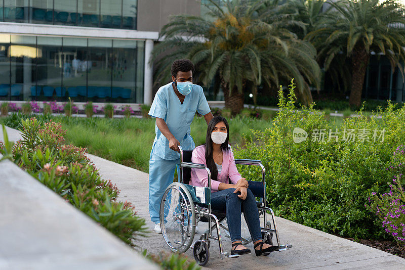 黑人男护士用轮椅抬着一位年轻的女病人穿过医院内花园。新常态。