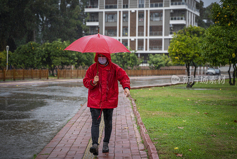 成熟的男人穿着红雨衣，撑着红伞走在雨中的城市街道上