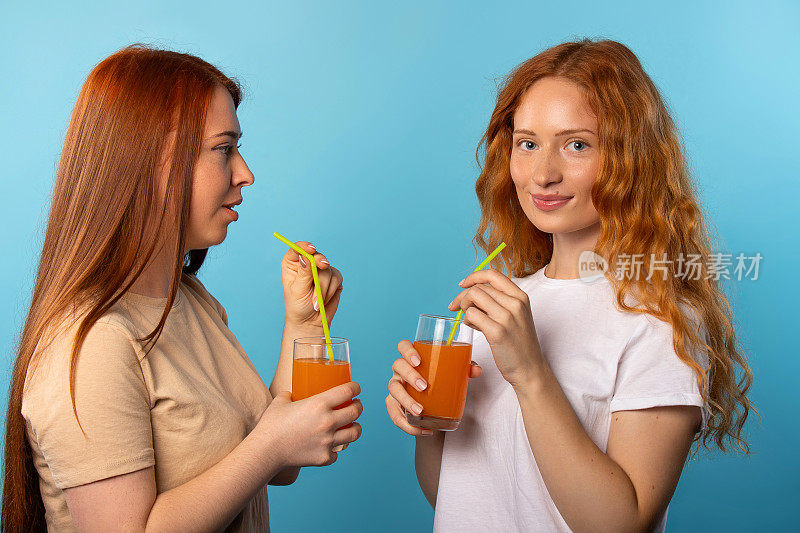 两个红头发的朋友边喝橙汁边看镜头。蓝色背景和自由时间概念。