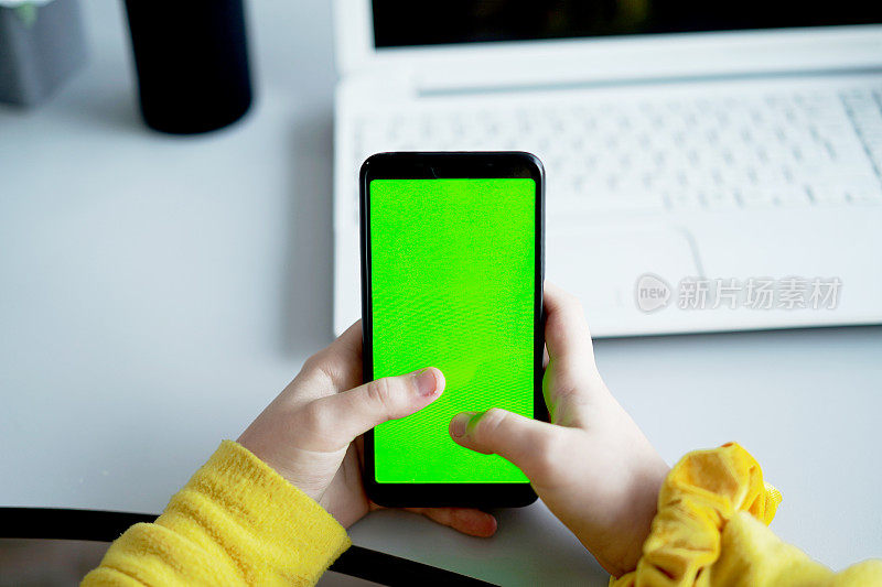 特写的十几岁的女孩手持智能手机与色度键绿色屏幕对白色笔记本电脑