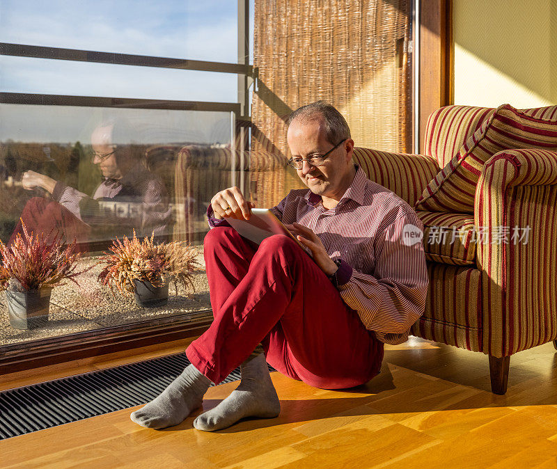 一位中年白人老人坐在家里的客厅里，靠在扶手椅上，享受着他的数码平板电脑