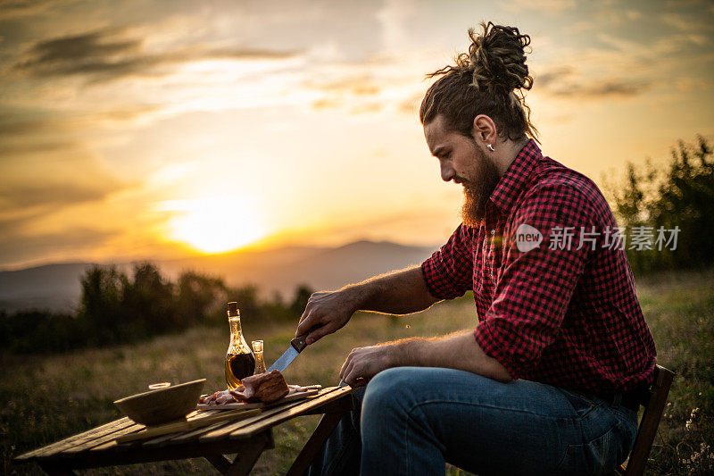 一个在山上的男人，一个伐木工人，喝着自制的白兰地，吃着自制的食物，而在他身后是美丽的日落，休息辛苦的工作。