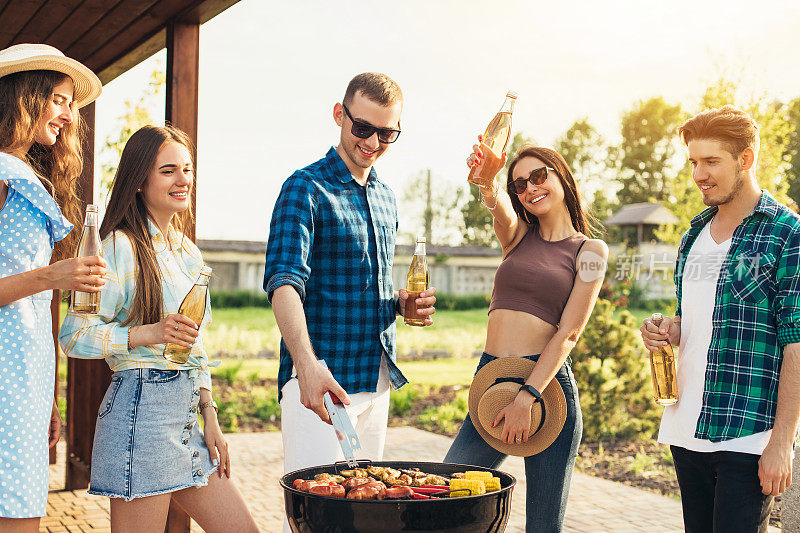 一名男子在夏季花园聚会上做烤肉，一群朋友散步、聊天、喝饮料