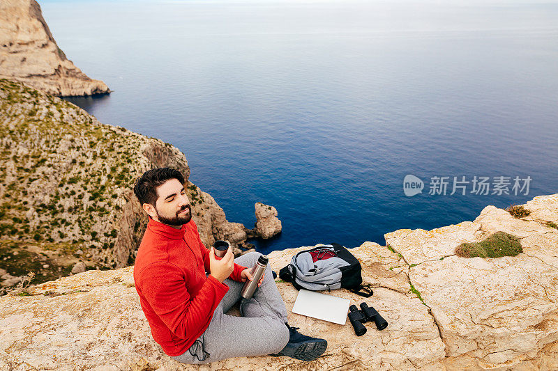 一个年轻的徒步旅行者坐在他的笔记本电脑，望远镜，背包上，闭着眼睛享受着从保温瓶热茶在石壁上，背景是马略卡岛北海岸的波伦萨湾的海洋