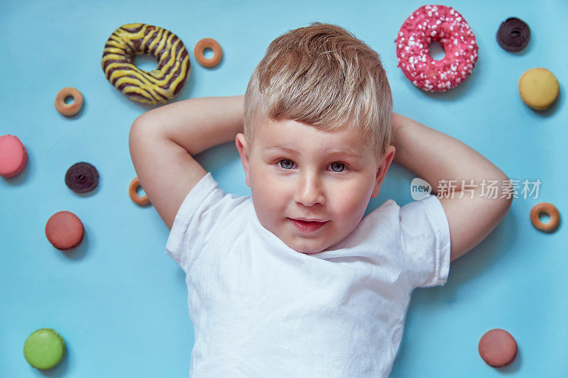 可爱的微笑的孩子甜甜圈和法国马卡龙在蓝色的背景。国家甜甜圈日概念。快乐的童年的概念。模拟白色t恤