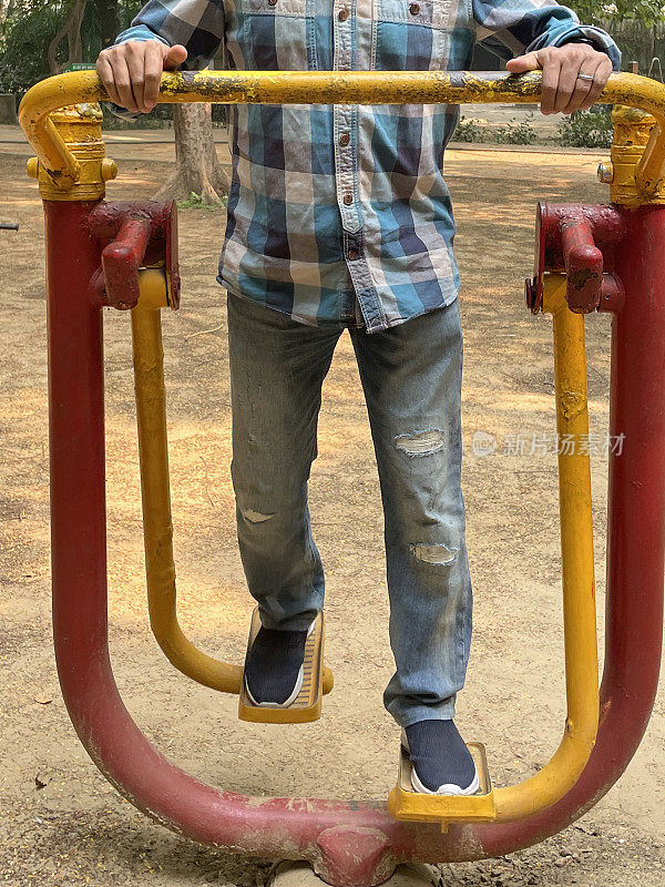 一个难以辨认的特写图像，印度男子在牛仔裤和衬衫在户外健身房使用空气行者健身运动设备，在户外露天公园锻炼