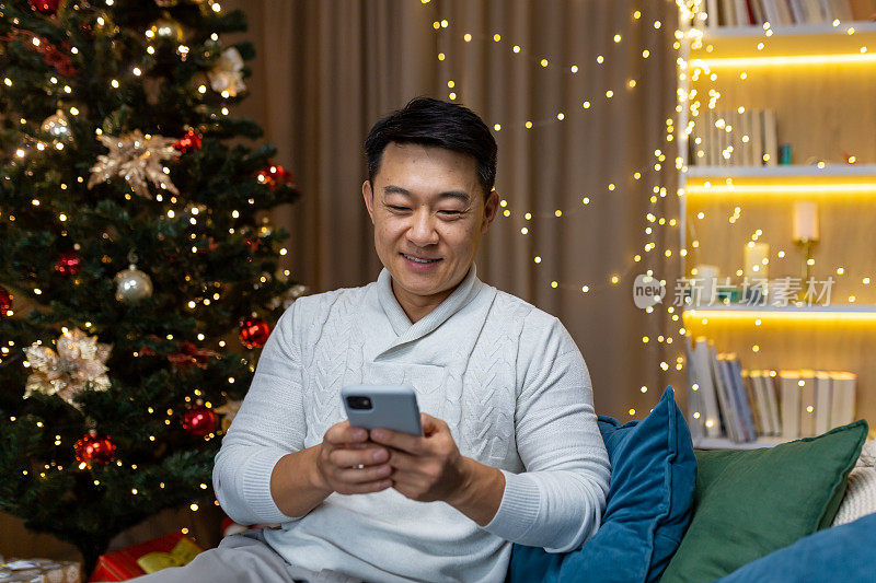 年轻英俊的亚洲男人坐在家里，靠近圣诞树。拿着手机，打字