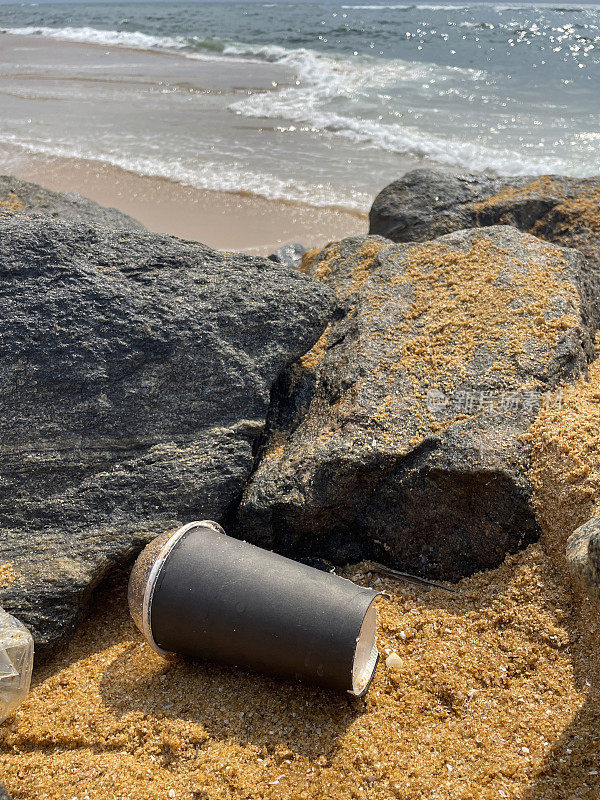 图像的空，纸板一次性咖啡杯与透明塑料圆顶盖冲在岩石池在退潮，海洋垃圾和污染的海岸，垃圾的沙子，肮脏的海滩，破碎的海浪背景，重点在前景