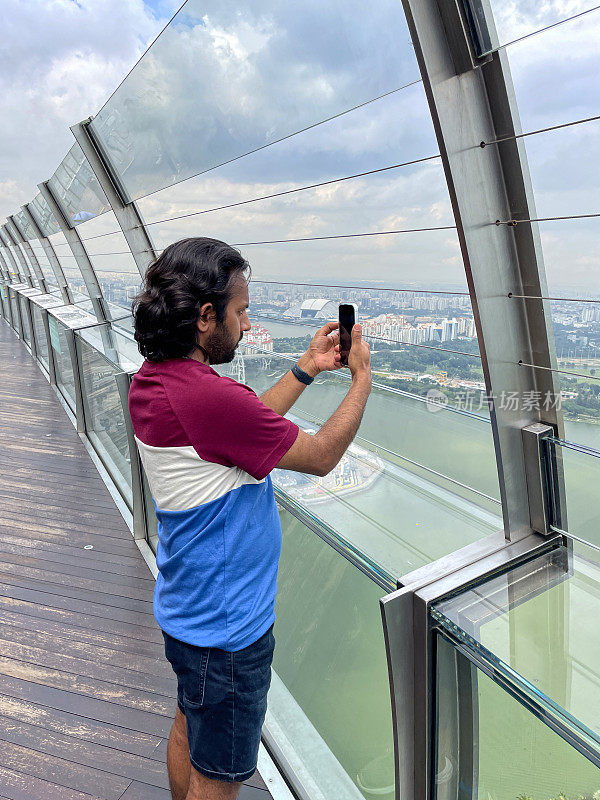 印度男子站在观景台安全屏障后的特写，拍摄市中心全景，背景是新加坡的城市景观摩天大楼，重点是前景