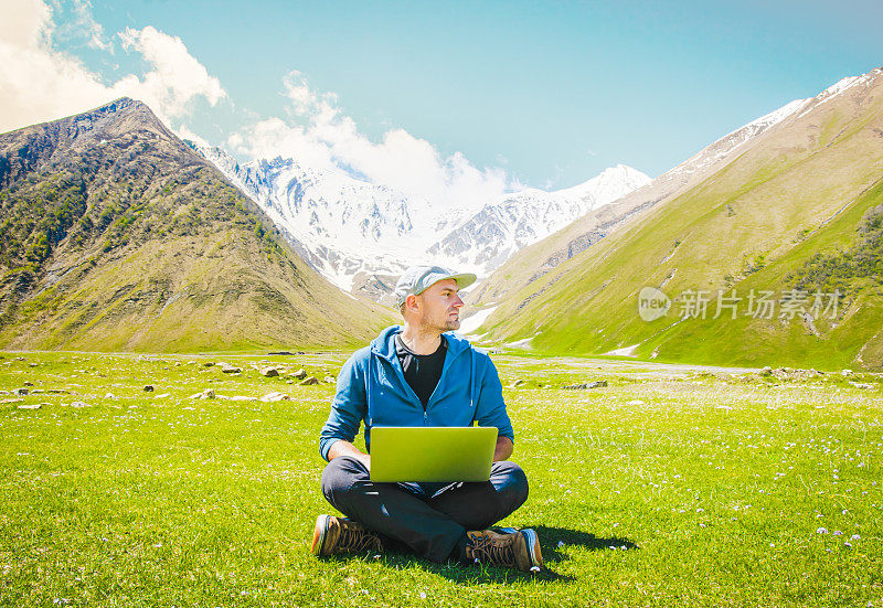 白人帅哥数字游牧民坐在笔记本电脑上的工作类型与山脉背景。数位游民旅行博客。大流行后世界的在线想法和工作。