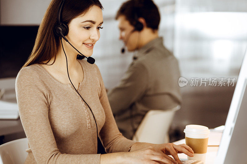 在一间现代化的办公室里，戴着耳机的年轻友好的女孩和她的同事坐在办公桌前，与公司的客户交谈。呼叫中心接线员正在工作