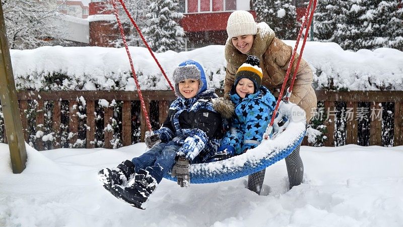 两个兄弟和妈妈在公共操场上愉快地玩着雪地秋千。冬季乐趣，家庭纽带，以及孩子们的户外活动。