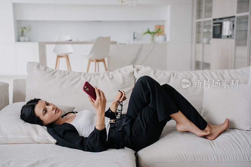 在现代客厅里，一位女商人懒洋洋地躺在沙发上玩智能手机