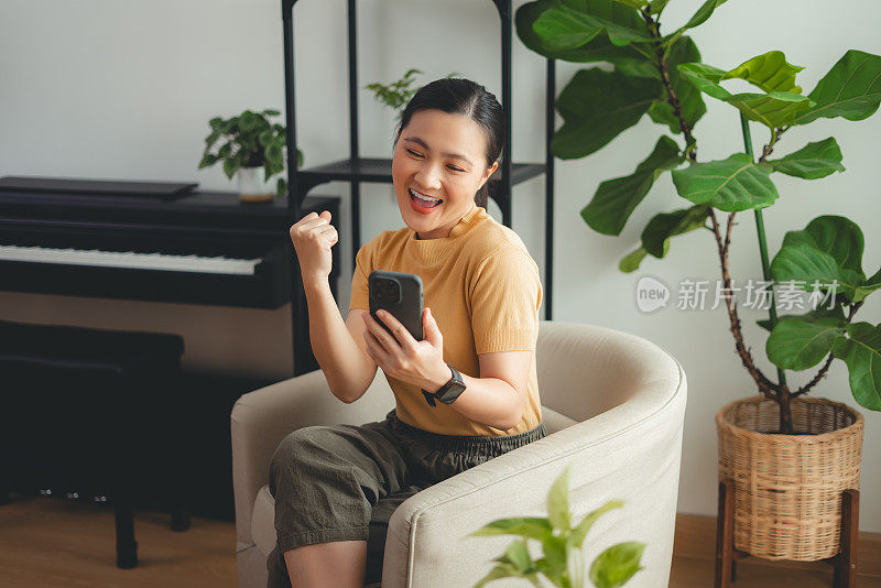 亚洲女性坐在家里的扶手椅上，用智能手机开心兴奋地做出了获胜的手势。