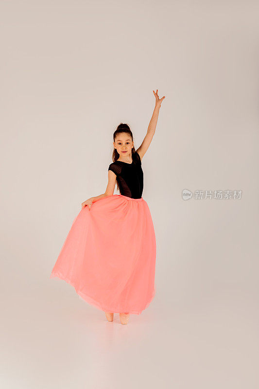芭蕾舞的概念。一个穿着黑色体操紧身衣的女孩，在白色的背景上，穿着尖头鞋和粉红色的裙子，摆出美丽优雅的姿势