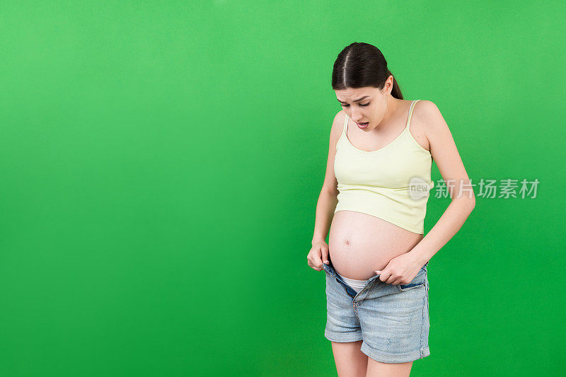 孕妇穿着拉开拉链的牛仔裤，在彩色的背景上露出赤裸的腹部。待产概念