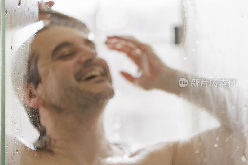 一个成熟，快乐的50岁男人在洗澡。脸是散焦的，焦点在淋浴门的湿玻璃上。