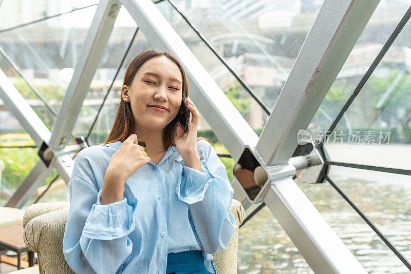 年轻聪明的亚洲女商人在蓝色寒意衬衫快乐地微笑在电话，当信用卡呼叫中心批准她的临时信用额度增加