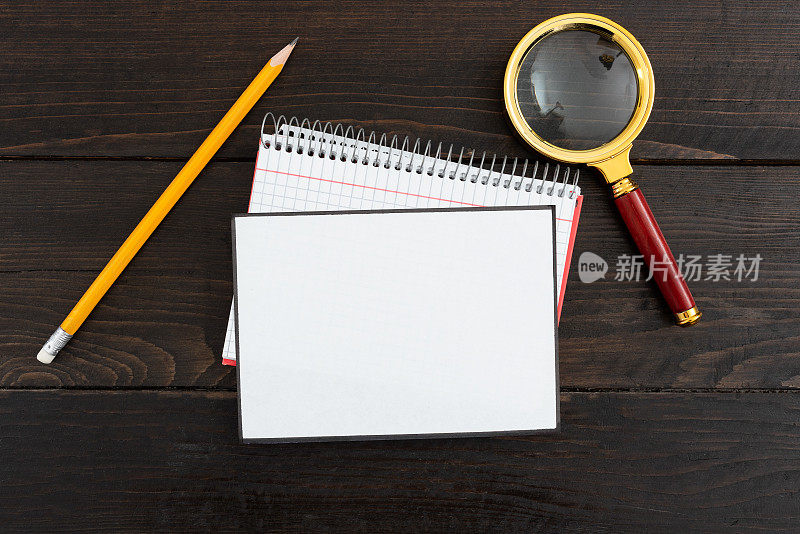 白纸与螺旋笔记本，铅笔和放大镜在木制背景。桌子上放有文具的空纸。它显示了重要的商业信息。