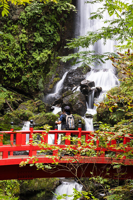 日本的女性游客在一座有瀑布的红色桥上驻足欣赏风景