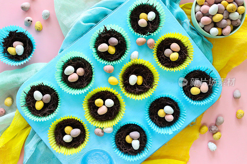 松饼烤盘上有一排排自制巧克力燕窝蛋糕，放在色彩鲜艳的纸蛋糕盒里，上面有迷你巧克力复活节彩蛋，蓬松的黄色小鸡玩具装饰，黄色，绿色和蓝绿色的棉布，粉红色的背景