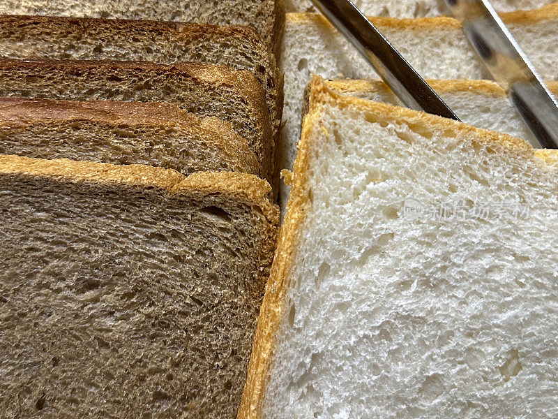 面包房货架上一排排的面包片堆叠在木托盘里的全帧图像，白色和全麦面包片，金属服务钳，重点在前景