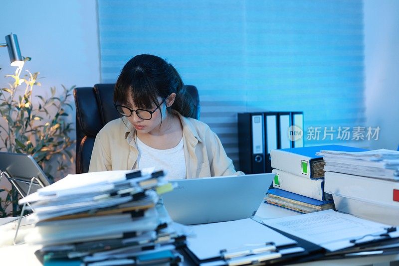 年轻的亚洲女商人在深夜工作。晚上，她坐在漆黑的家庭办公室里，忙着检查文件，不得不在截止日期前把工作送出去。