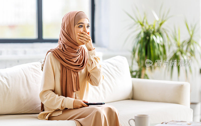 震惊的穆斯林妇女戴着头巾，拿着电视遥控器在家看电视剧