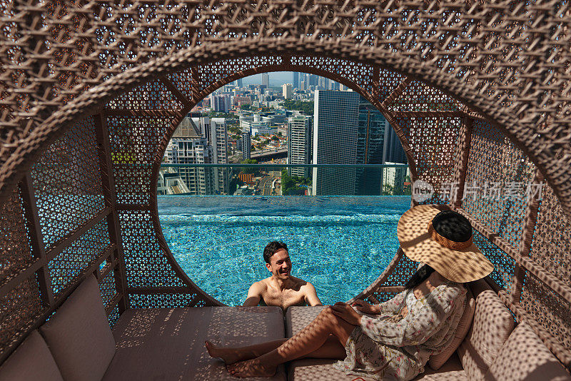 一对度假的亚洲夫妇从酒店的屋顶无边泳池欣赏城市天际线